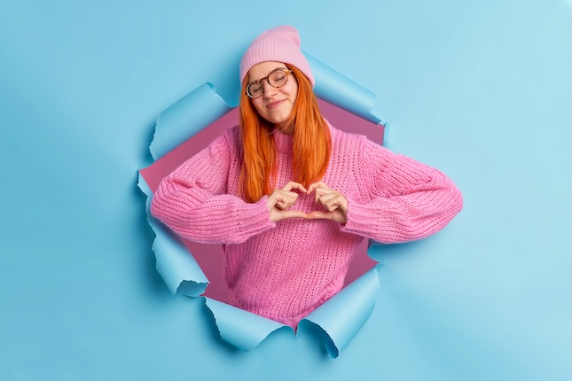 romantic-concept-satisfied-affectionate-redhead-woman-does-heart-symbol-love-sign-shapes-heart-with-fingers-closes-eyes-with-pleasure-wears-pink-hat-sweater-breaks-through-paper-wall
